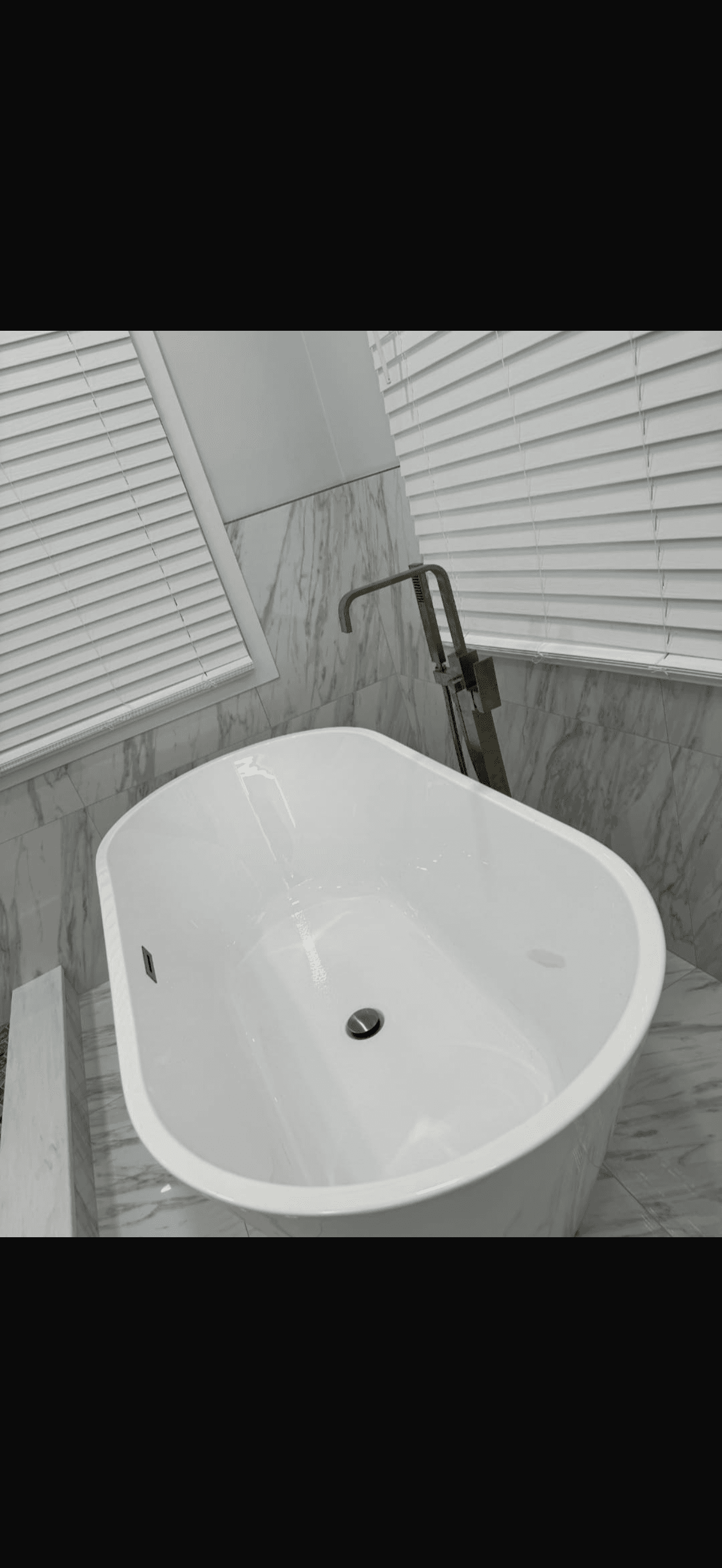 Weiße ovale freistehende Badewanne vor Marmorwänden mit einer modernen, hohen Bodenarmatur aus gebürstetem Metall.